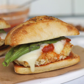 Baked chicken parm sub on mini baguette bread. One sandwich is in focus with brown sandwich paper on the bottom. Two sandwiches are out of focus and fresh basil pieces added for styling purposes.