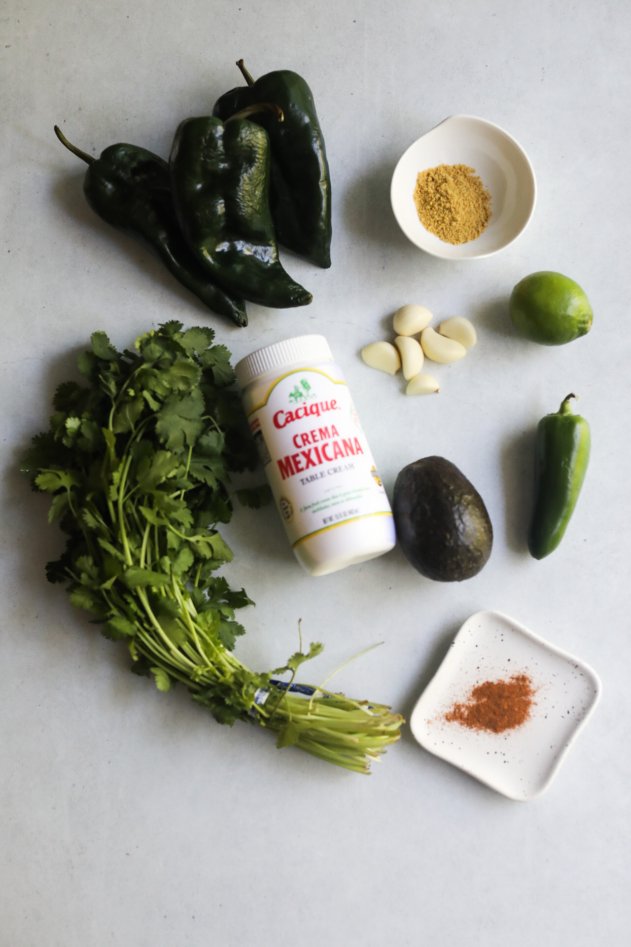 Poblano Cream Sauce ingredients on a flat lay with cilantro, poblano peppers, Mexican crema, chicken bouillon, fresh garlic, lime, avocado, jalapeno and cayenne pepper.