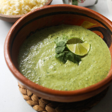 Poblano Cream Sauce (Culichi Salsa) in a clay/brown bowl topped with cilantro and lime wedges. Bowl of cheese and a white bowl of shredded chicken added for styling purposes.