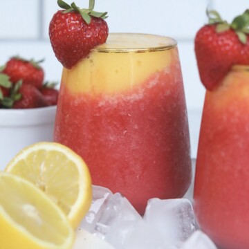 Wine slushy poured in a wine glass. Frozen strawberry and mango slushy in a wine glass. White bowl with strawberries and a flat white plate added with a sliced lemon and crushed ice.