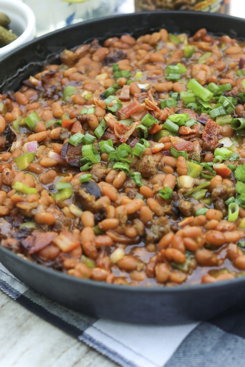 Baked Bean Casserole with Sausage in a black cast iron skillet. Small bowl with small pickles and black and white towel added for styling purposes.