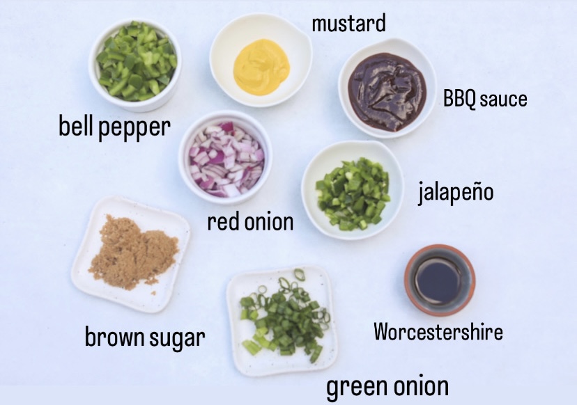 Sausage and Baked Bean Casserole ingredients in a flat lay. In small white bowls are green bell peppers, mustard, BBQ sauce, red onion, jalapeno, brown sugar, green onion and in small brown and blue bowl is Worcestershire sauce.