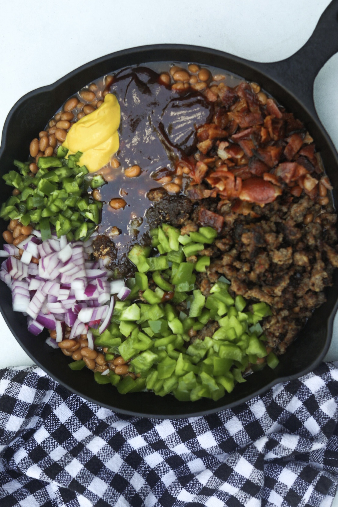 Baked bean casserole in a cast iron skillet with ingredients layered on top of the baked beans. Chopped jalapeno, red onion, green bell pepper, mustard, BBQ sauce, ground sausage and crumbled bacon.