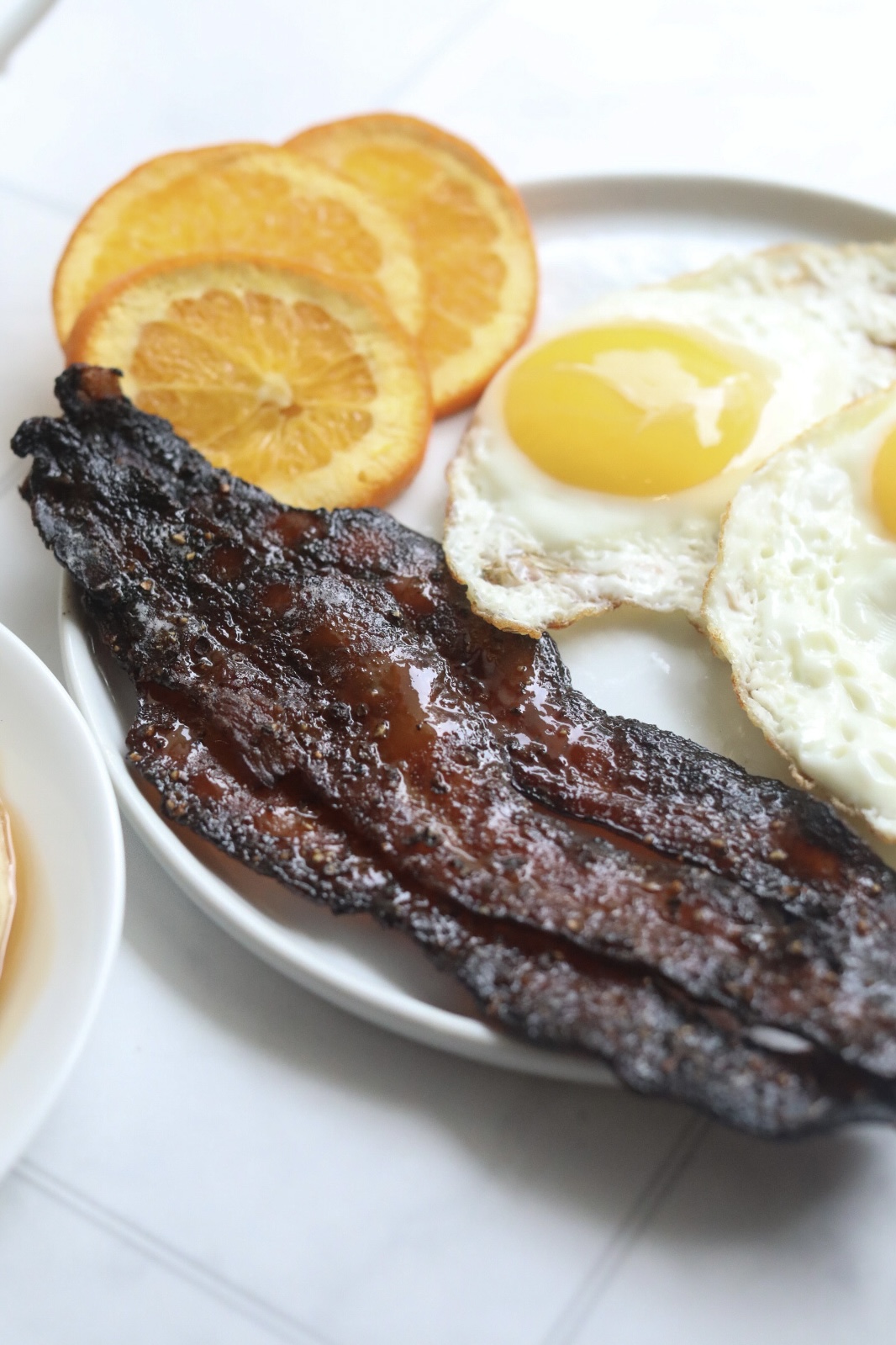 Maple candied bacon on a white plate with fried eggs and sliced oranges.
