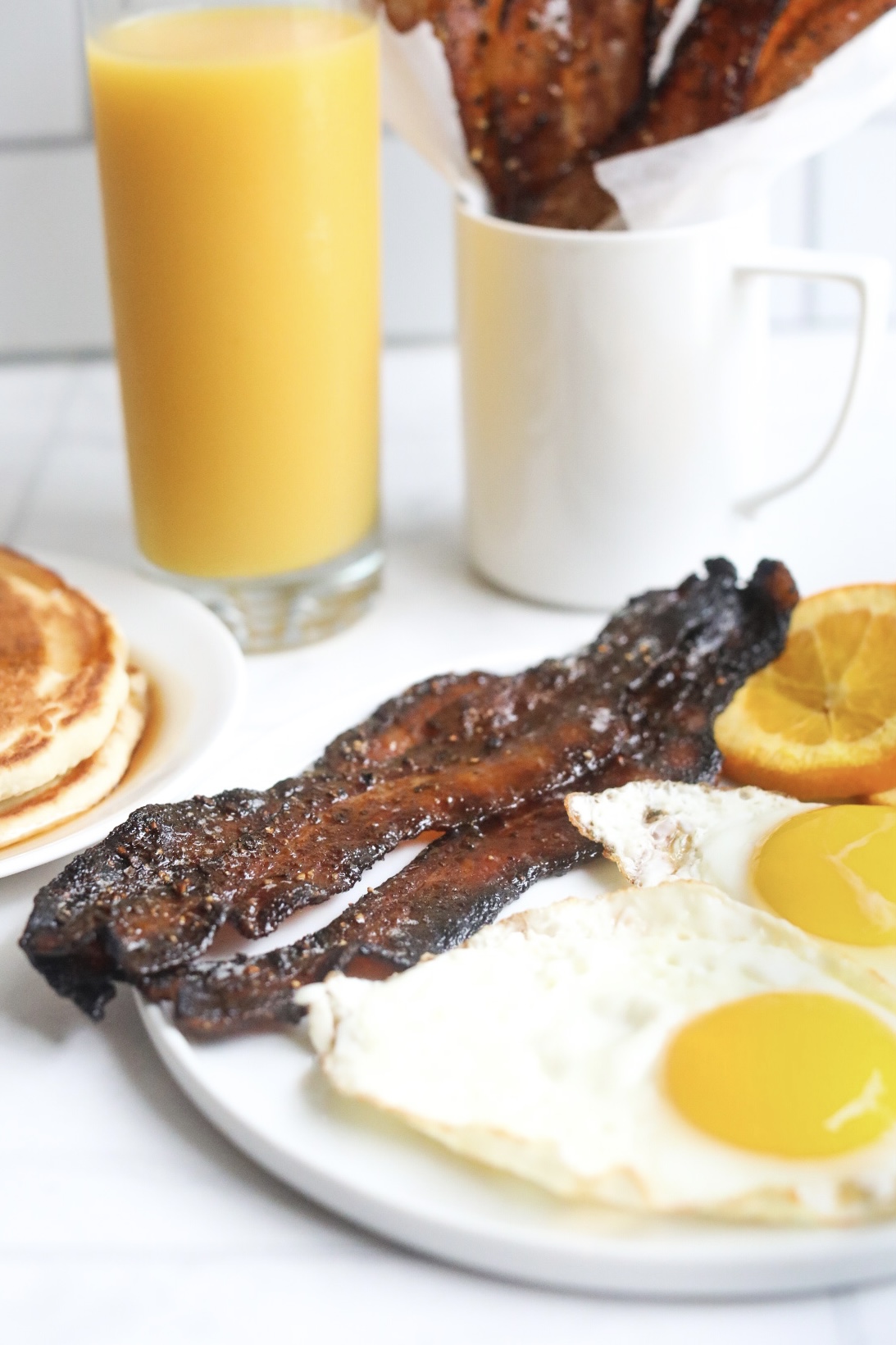 Candied bacon on a plate with breakfast food. Suny side up eggs, sliced oranges and candied bacon. Out of focus and for styling purposes is a glass of orange juice, candied bacon in a mug and a small plate of pancakes.