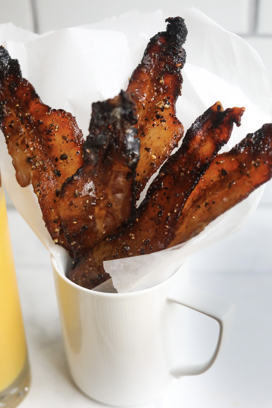 Candied bacon in white coffee mug, stuffed with white parchment paper. Glass of orange juice is slightly shown for styling purposes.