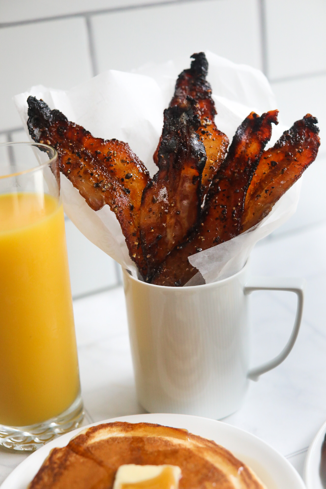 Maple candied bacon in white coffee cup with parchment paper lining the bacon. A glass of orange juice and a small plate of pancakes with butter on top is shown for styling purposes.