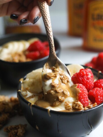 Homemade whipped yogurt served in a black bowl, topped with raspberries, granola and nut butter. Gold spoon is scooping out contents with hand that has black nails.