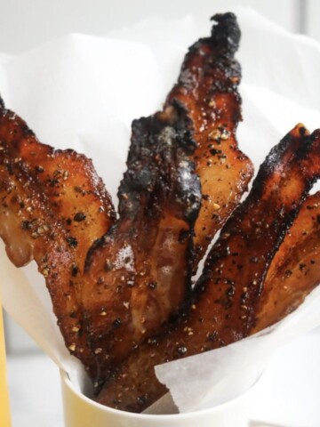 Maple candied bacon in a white coffee mug with a glass of orange juice on the side.