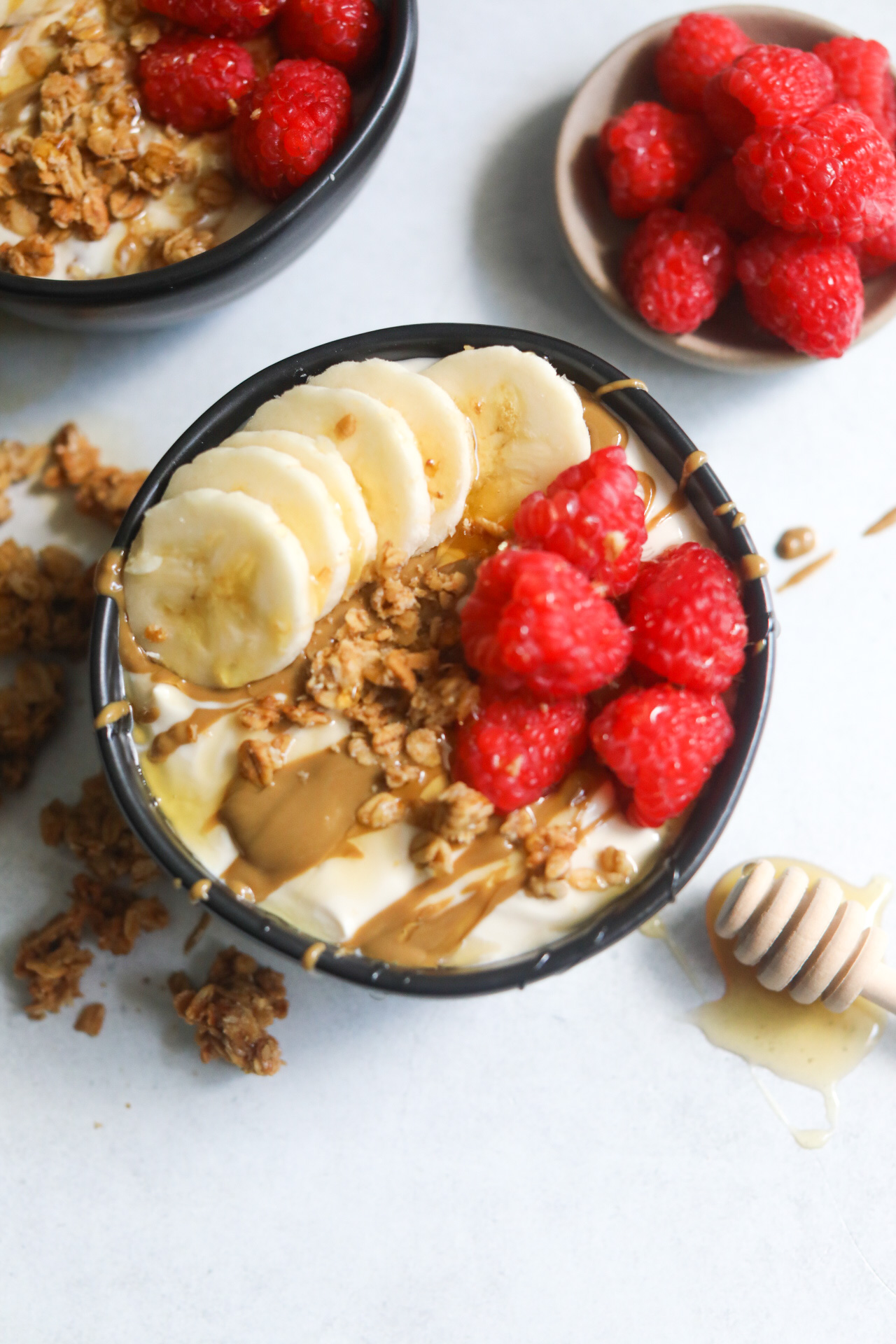 Whipped yogurt in a black bowl topped with sliced bananas, raspberries, granola and drizzled honey and sunflower butter. One bowl is centered and off centered is the corner of another dressed bowl with a small bowl of raspberres to the right in a small bowl.