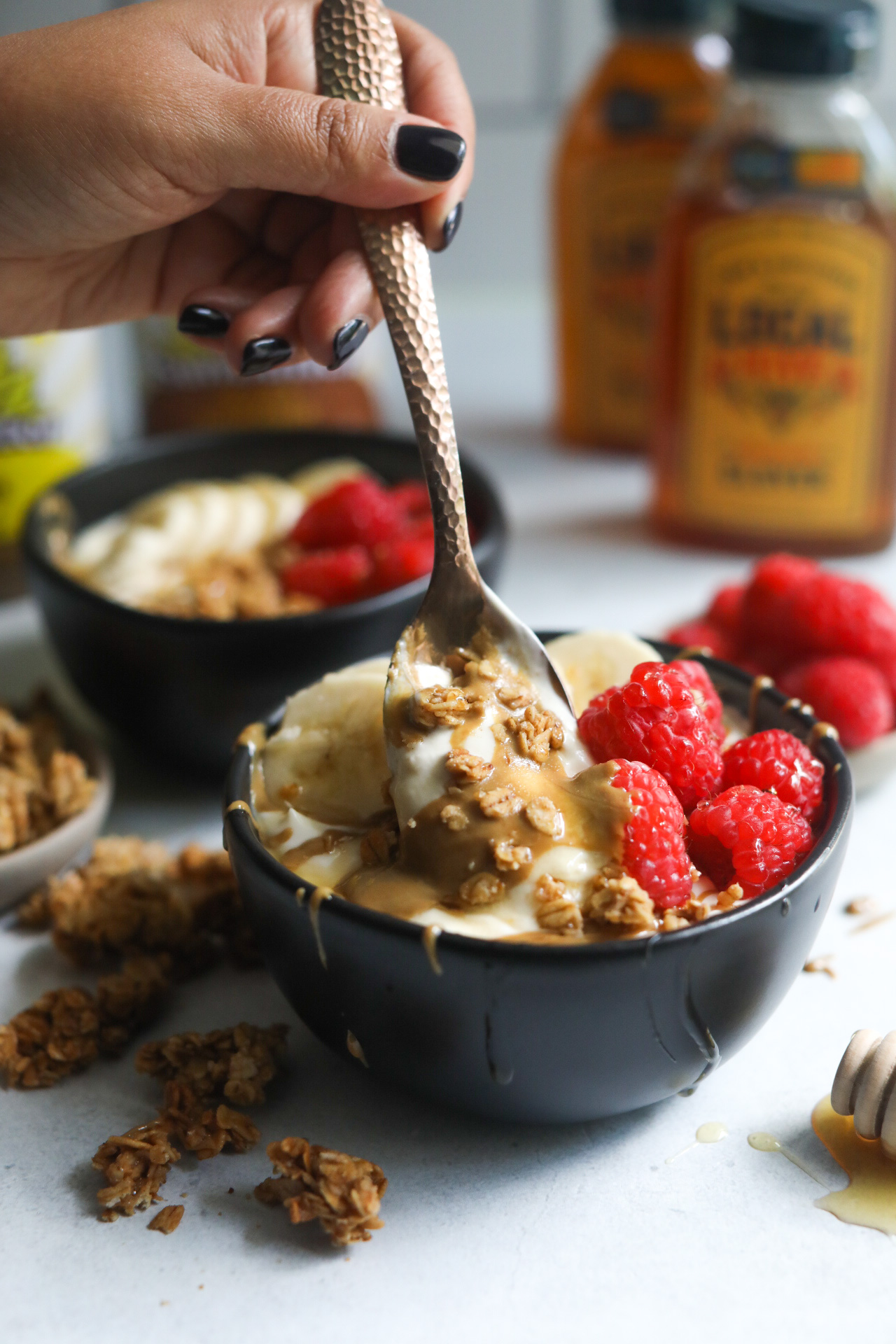 Whipped Greek yogurt bowl dressed with toppings (sunflower butter, sliced bananas, raspberries and granola) with a bronze decorative spoon with a hand scooping yogurt content out.