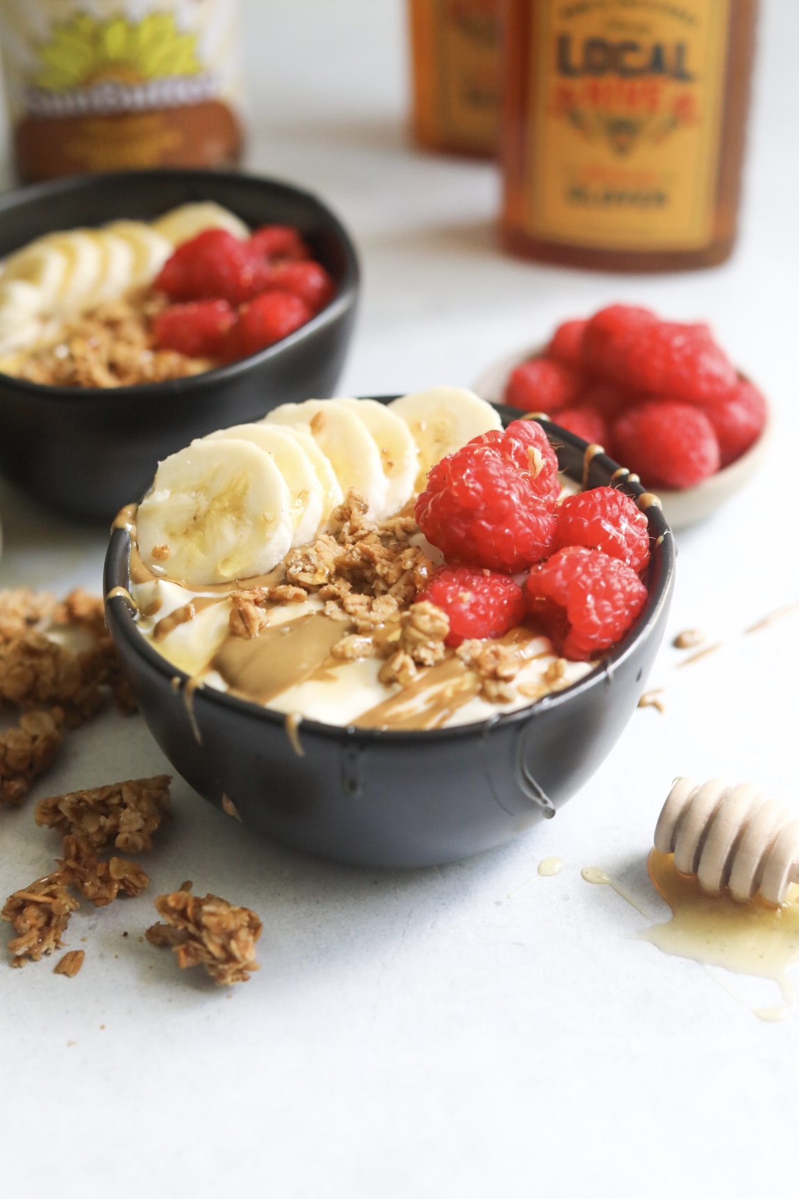 Whipped yogurt bowl with toppings in black bowl with a honey stick to the left of the bowl for decorative purposes. Out of focus behind the focal bowl is an additional dressed bowl with bottles of honey and sunflower butter.