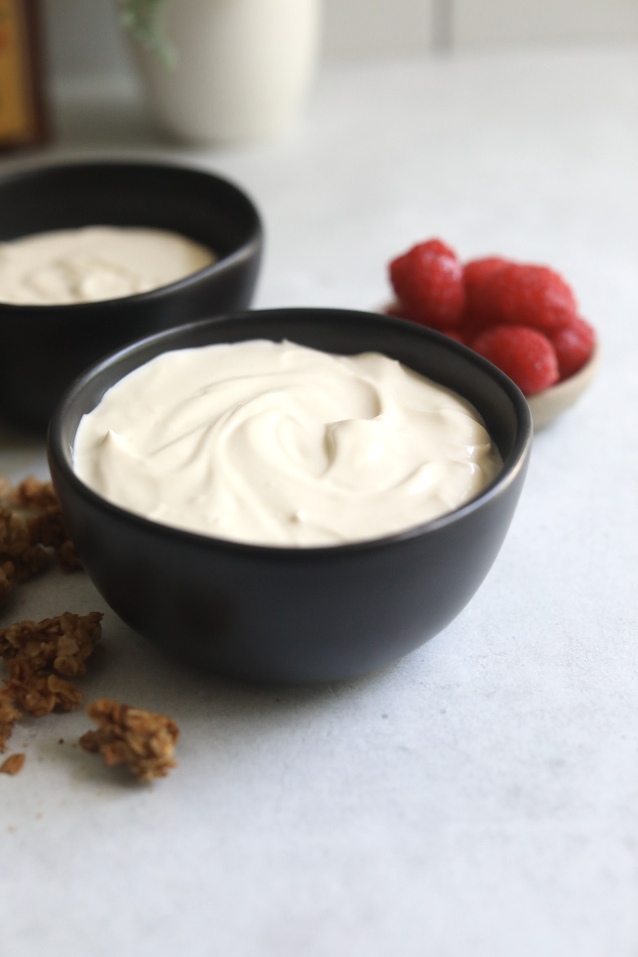 Whipped yogurt in a black bowl shocasing final recipe without toppings. Out of focus is an additional bowl with whipped yogurt and a small bowl of raspberries for styling purposes.