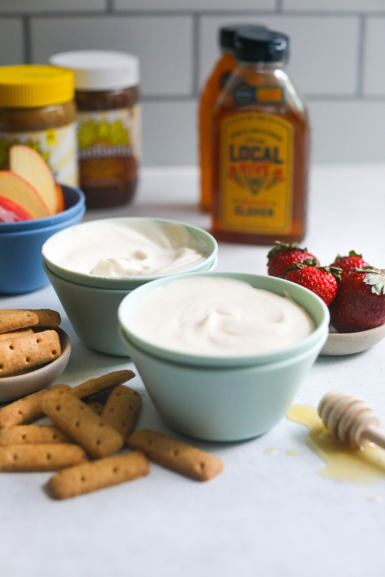 Whipped yogurt in kid friendly bowls (in the color light teal), with graham cracker sticks, sliced apples, small bowl of strawberries added and a honey stick with drizzled honey added for styling purposes.