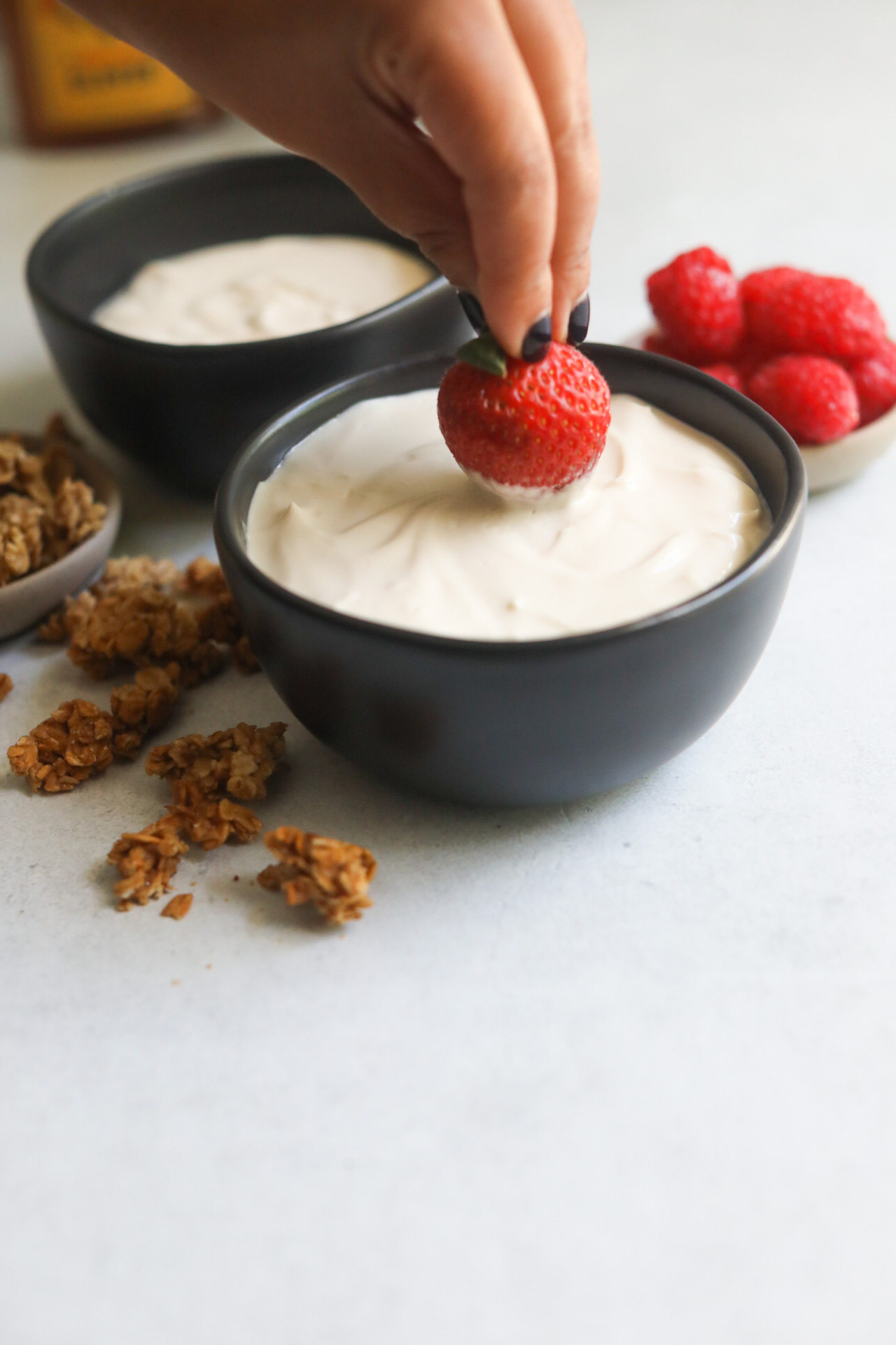 Hand dipping strawberry entirely into whipped yogurt bowl. Bowl is black with crumbled granola and a small bowl of raspberries added for styling purposes.
