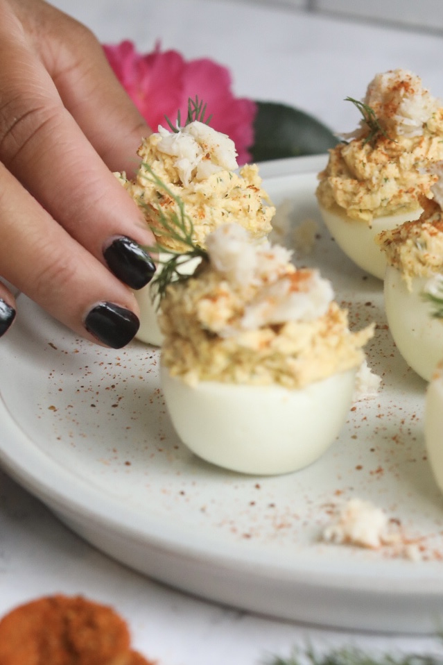 a hand reaching for a crab deviled egg from a platter