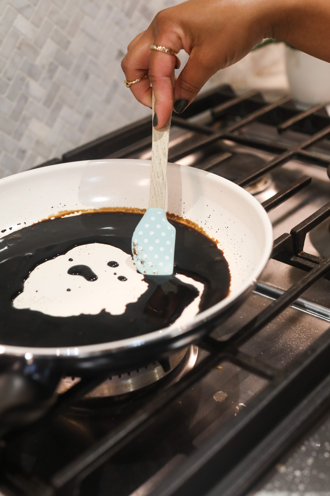 White and black pan on gas stove with balsamic vinegar thickening in pan with hand and a small spatula.