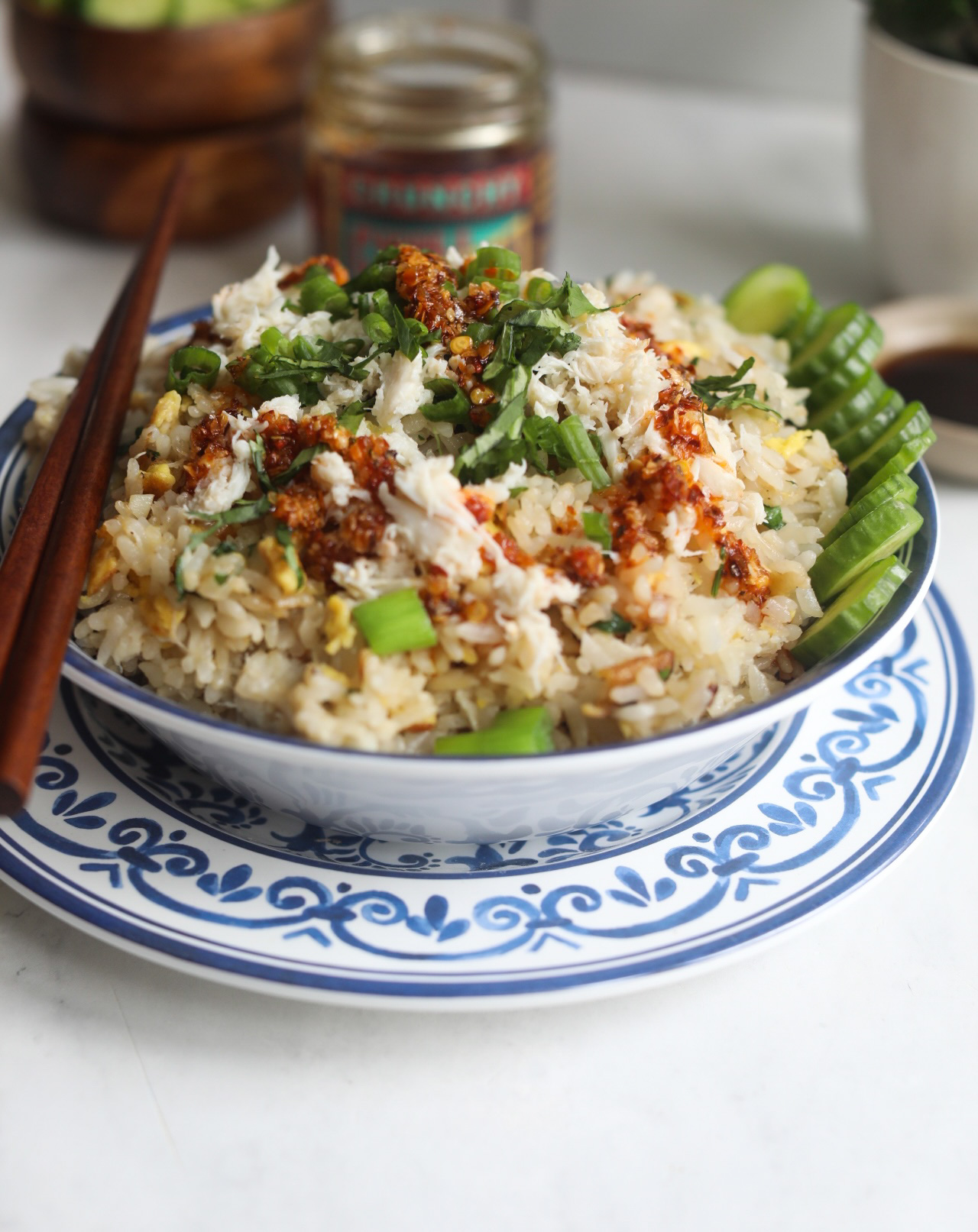 Crab fried rice in a blue and white decorative bowl garnished with sliced cucumber, green onion and cilantro. Brown chopsticks on the left side of the bowl for garnish.