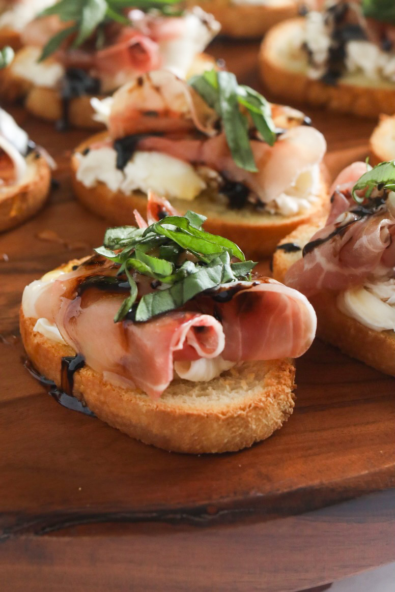 Close up of final recipe. Italian toast with burrata, prosciutto, balsamic glaze, honey and fresh basil. Toast points are on a wooden cutting board.