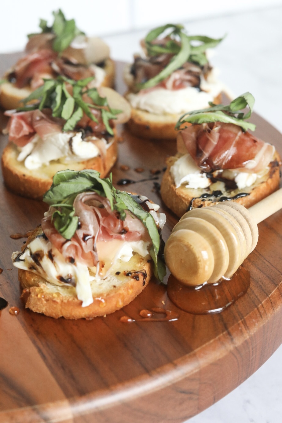 Italian toast with burrata, prosciutto, balsamic glaze, honey and fresh basil. Toast points are on a wooden cutting board. Honey dipper added for styling purposes.