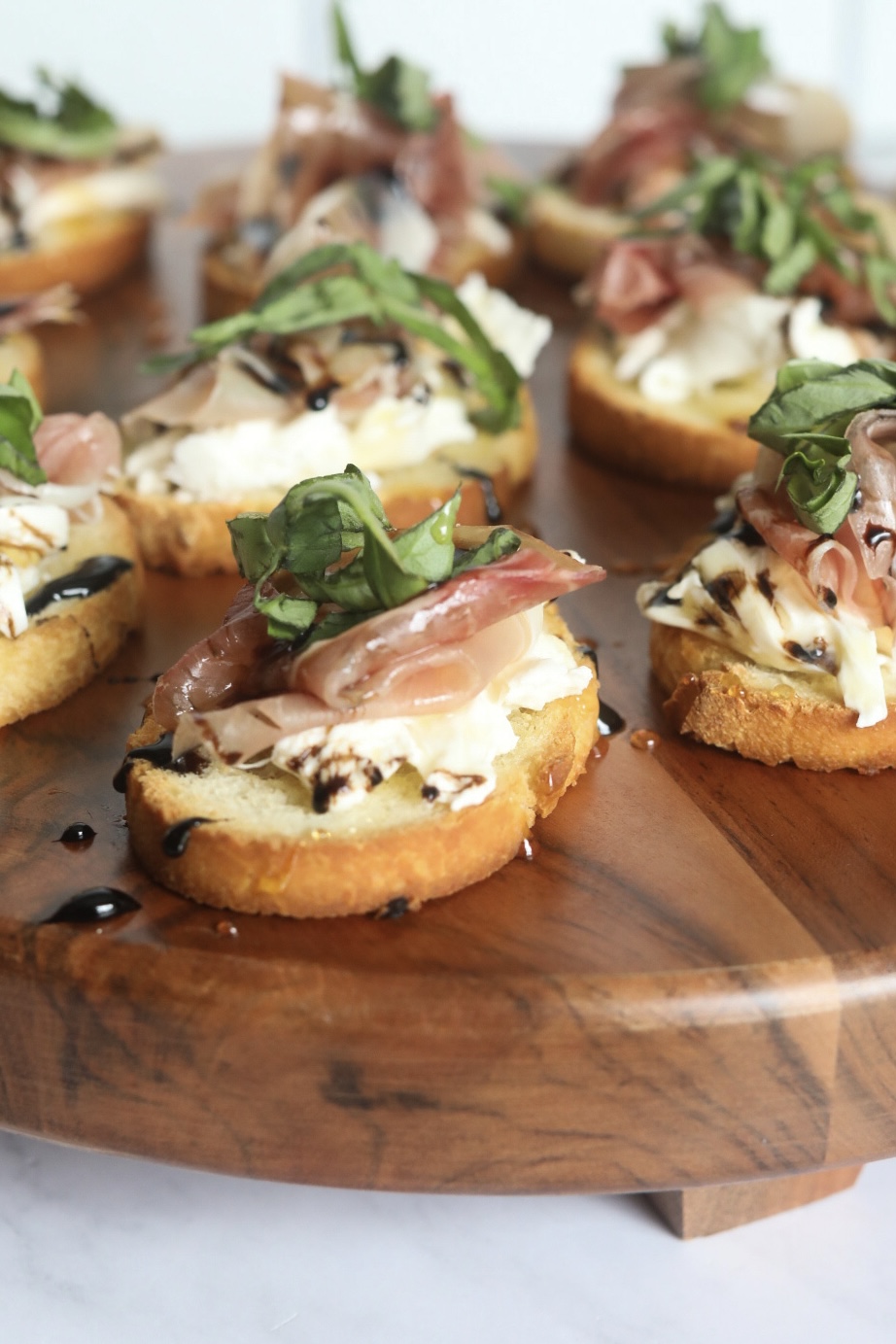 Italian toast with burrata, prosciutto, balsamic glaze, honey and fresh basil. Toast points are on a wooden cutting board.