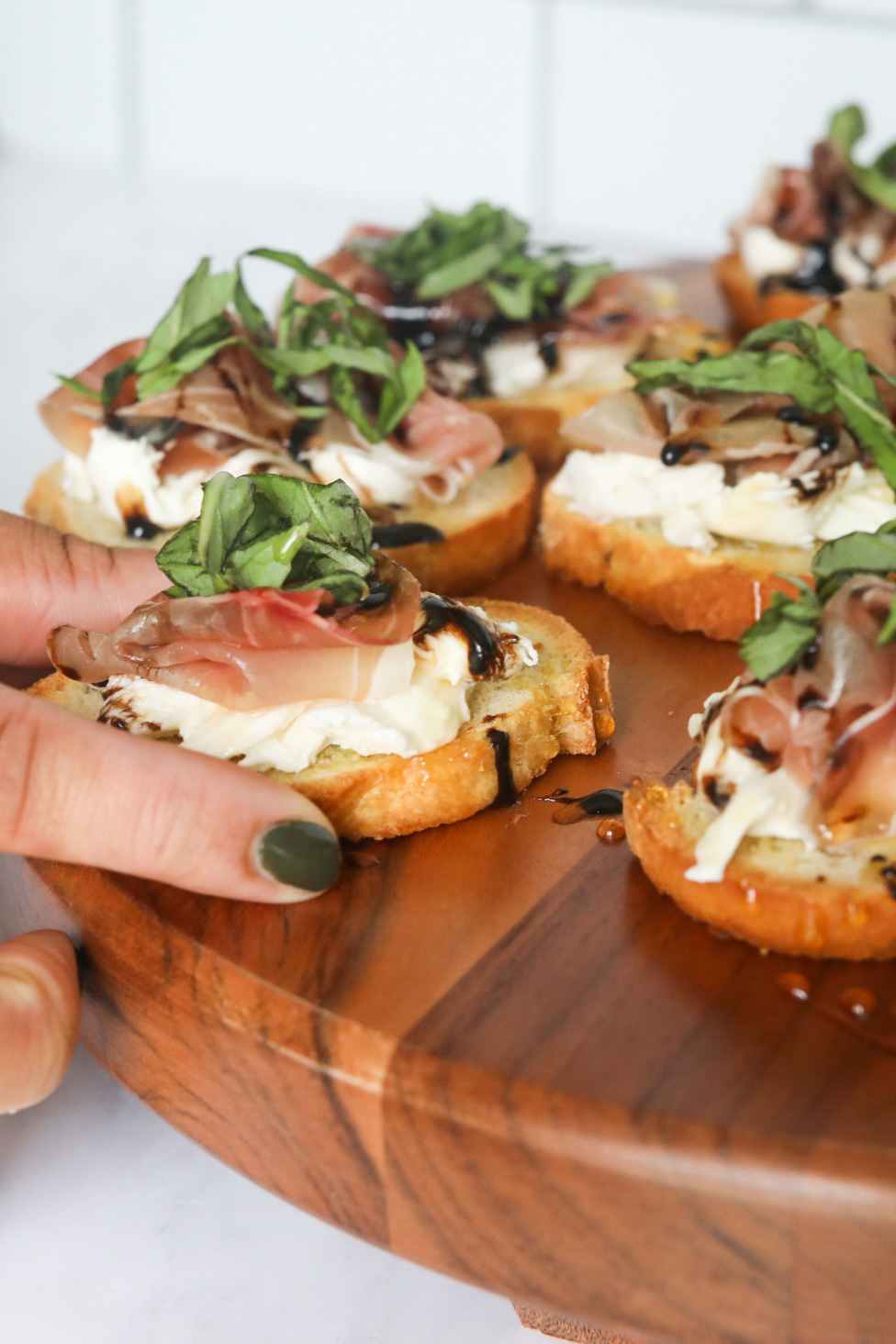 Hand picking up prosciutto crostini staged on a wooden charcuterie style board. Angle is slightly on the left-hand side with the hand picking up the toast point.