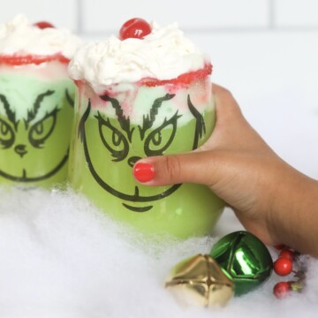 Small, toddler hand holding Grinch Punch. Clear stem-less cups are decorated with black stencil grinch faces. Green punch is topped with whipped cream and cherries. Decor is added for styling purposes, jingle bells and fake snow.