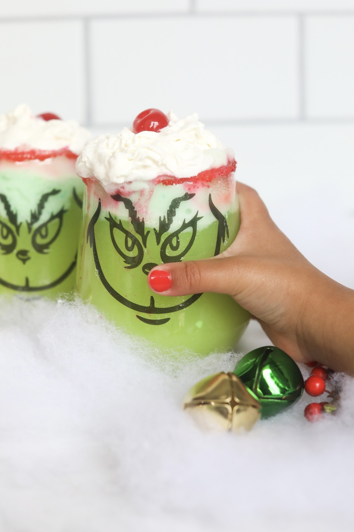 Small, toddler hand holding Grinch Punch. Clear stem-less cups are decorated with black stencil grinch faces. Green punch is topped with whipped cream and cherries. Decor is added for styling purposes, jingle bells and fake snow.