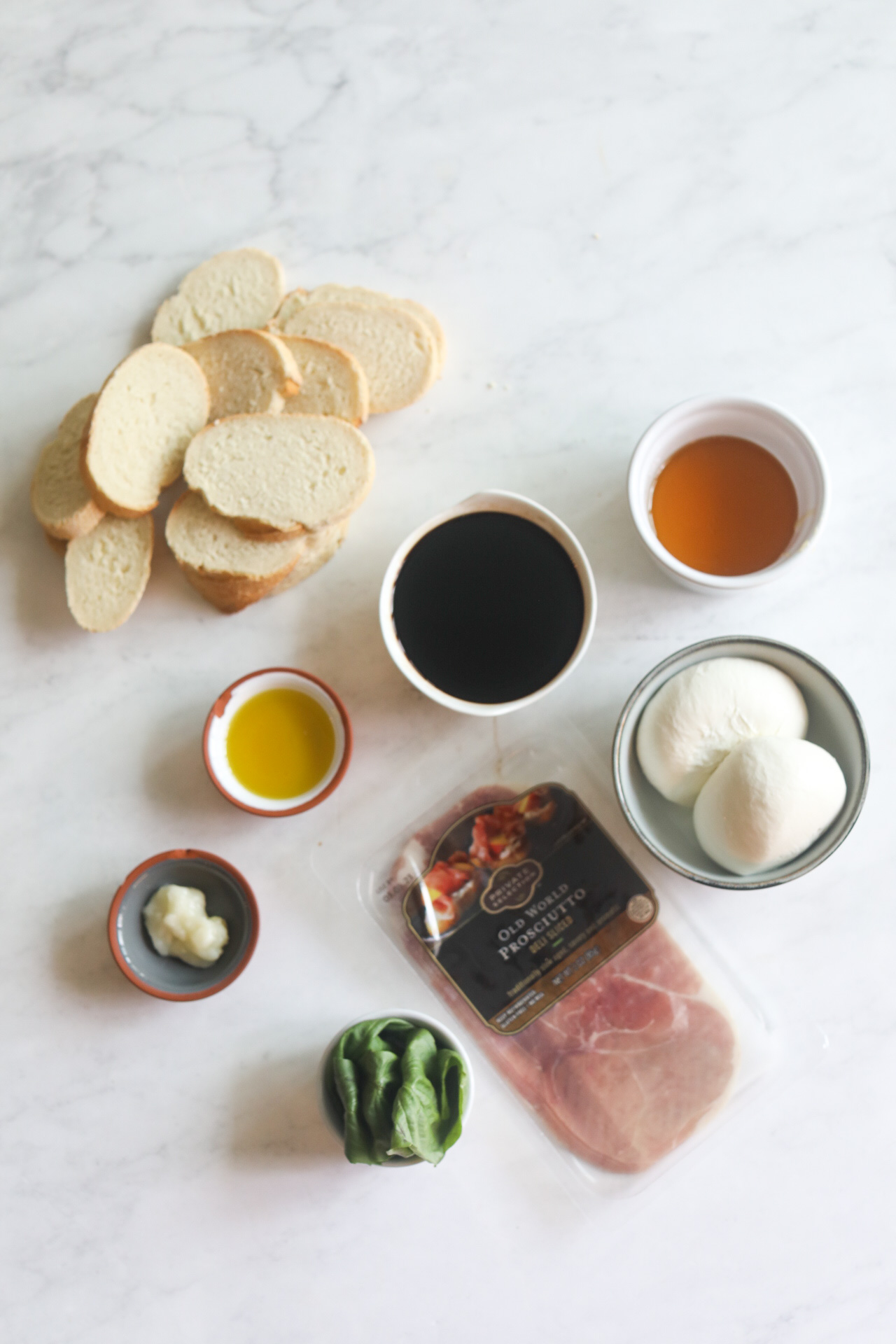 Prosciutto crostini with balsamic glaze and honey drizzle ingredients on a flat lay. Sliced Italian bread, small bowls of garlic, honey, balsamic, olive oil, prosciutto and burrata.