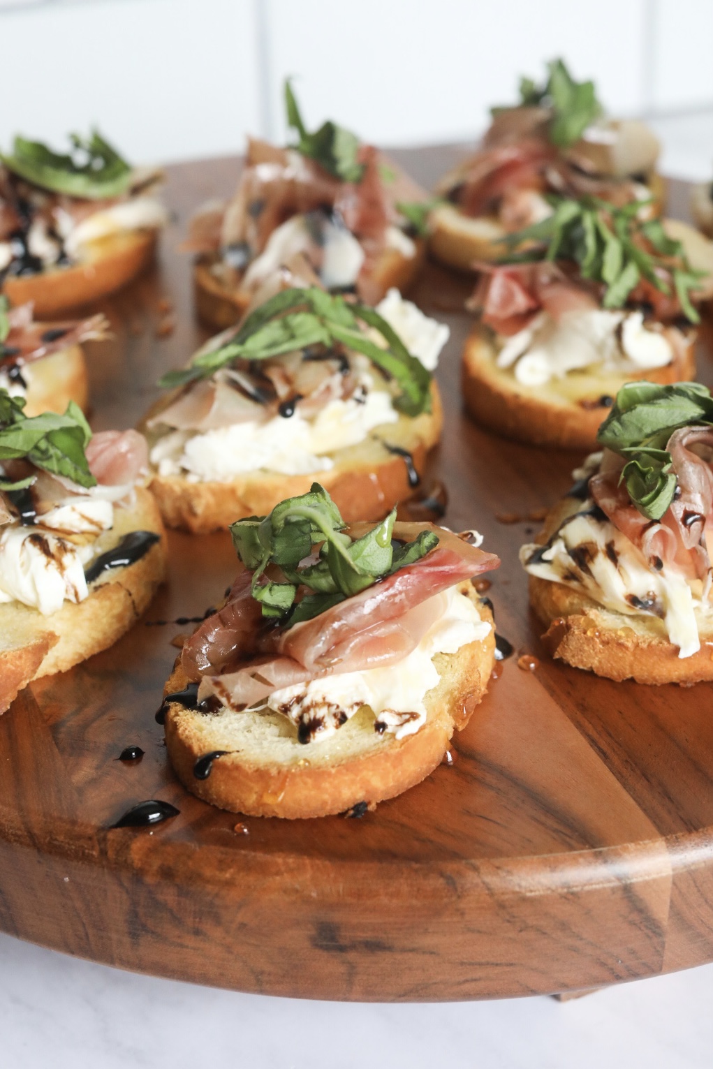 Crostini toast point with burrata cheese, prosciutto, fresh basil drizzled with balsamic glaze and honey on a wooden charcuterie board.