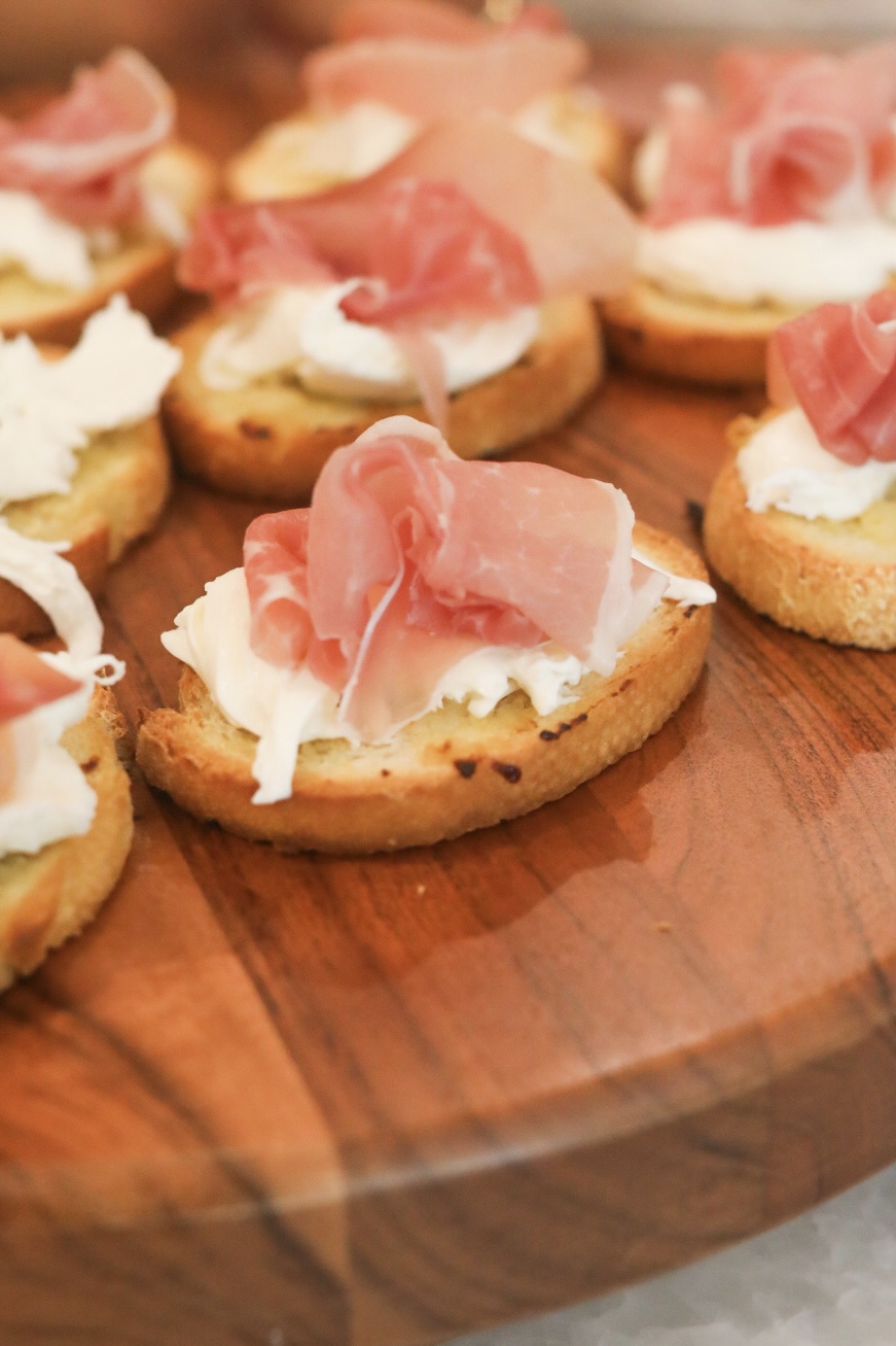 Toast point with toasted Italian style bread with burrata and prosciutto on a wooden charcuterie style board.