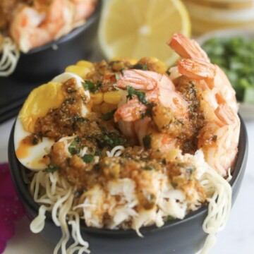 Spicy cajun seafood ramen with noodles, shrimp, crab, boiled egg and corn in a black bowl. Sliced lemon and a small bowl of green onions added for styling purposes.