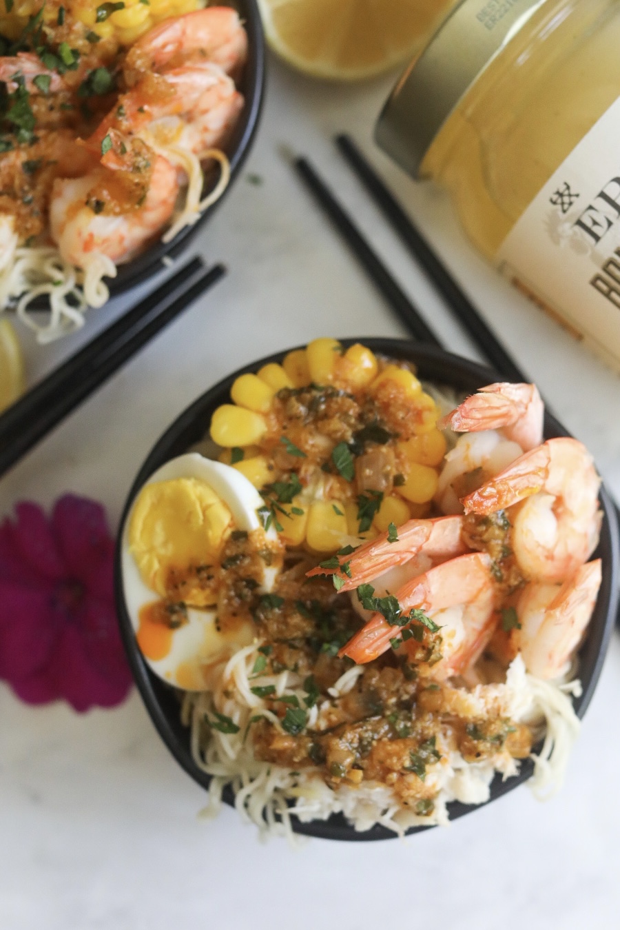 Top down view of finalized recipe, spicy cajun seafood ramen. Two bowls, with one in the center, focused filled with ramen, shrimp, crab, boiled eggs and corn. Cajun butter sauce is drizzled over bowl with fresh parsley.