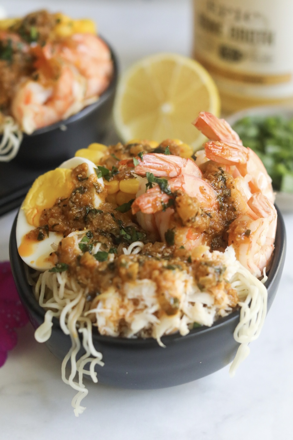 Spicy cajun seafood ramen with noodles, shrimp, crab, boiled egg and corn in a black bowl. Sliced lemon and a small bowl of green onions added for styling purposes. 