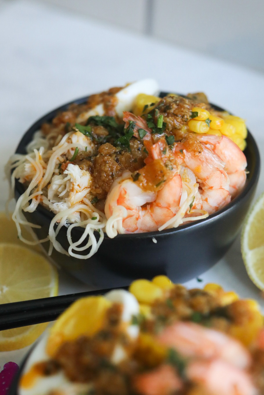 Zoomed in image of the cajun seafood ramen plated in a black bowl with sliced lemons and black chopsticks added for styling purposes.