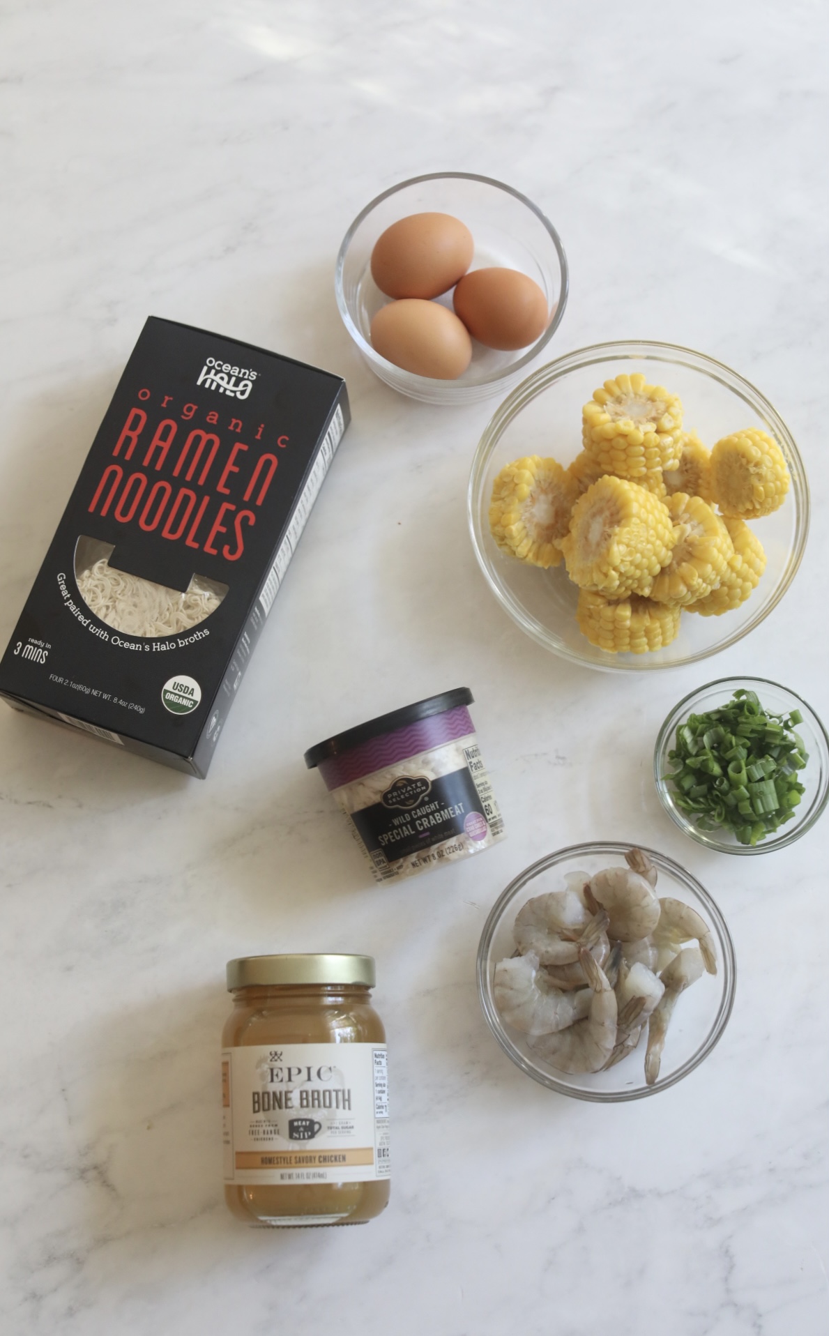 Seafood ramen ingredients in a flatlay. Black box of store-bought ramen noodles, jar of bone broth, can of crab meat and clear bowls of eggs, corn, green onion and shrimp.