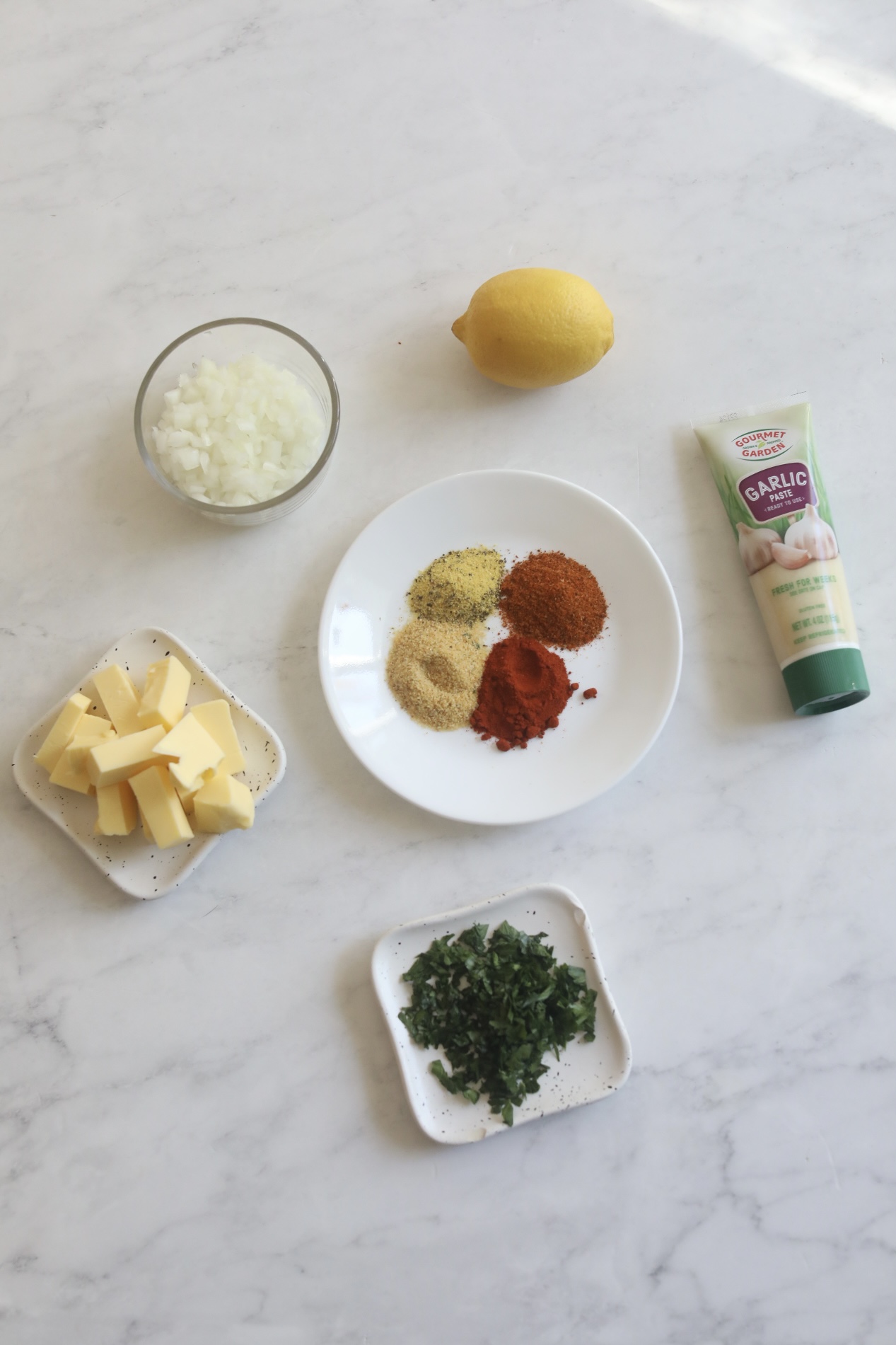 Spicy cajun butter sauce for seafood ramen. In flatay style, small white plate with seasonings, small white plates of sliced butter and parsley. Small clear bowl of onion, lemon and a tube of garlic paste.