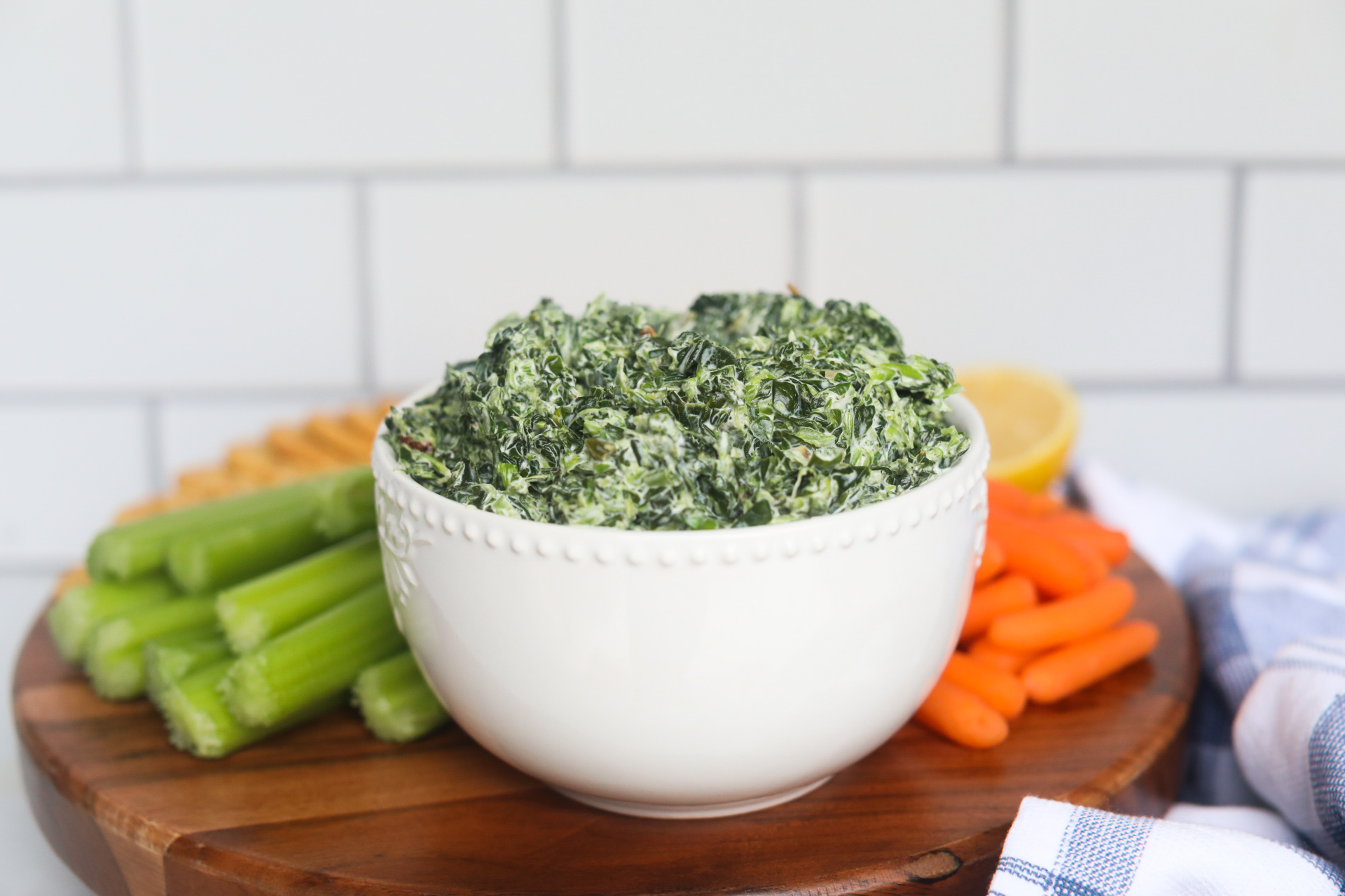 Spinach dip served in a white bowl with carrots, celery and crackers on a wooden circle board.