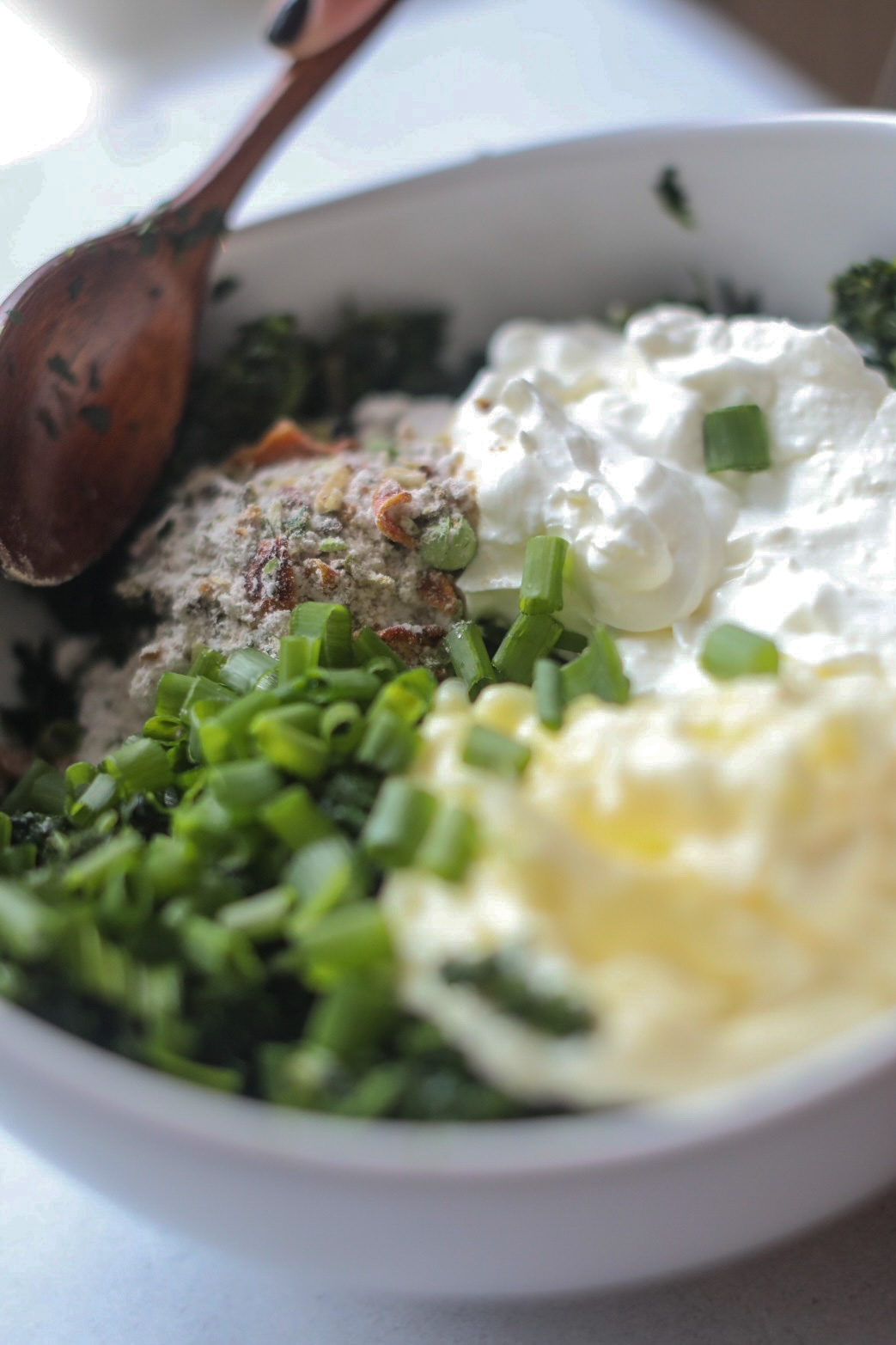 Cold spinach dip ingredients (spinach, mayo, sour cream, green onions, dried vegetable mix) in a white bowl with a brown wooden spoon.