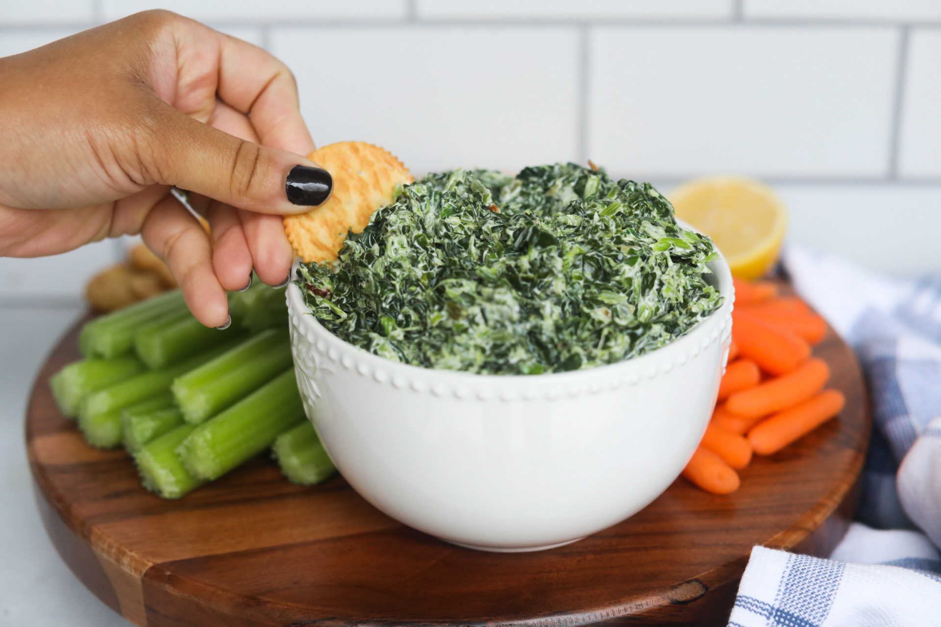 Hand dipping cold spinach dip with cracker out of the white bowl. Spinach dip is served on top of a wooden board with carrots, celery and crackers.