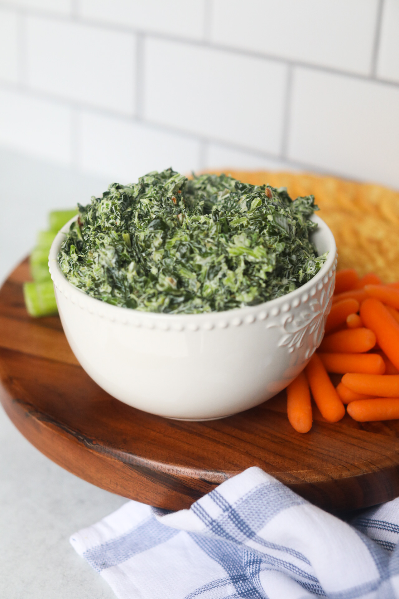 Complete and plated recipe served a white bowl on a brown wooden board. Surrounding the bowl of spinach dip with carrots, celery and crackers.
