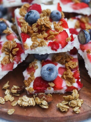 Yogurt bark cut into large squares served on a wooden cutting board. Granola is added around the yogurt bark for styling purposes.