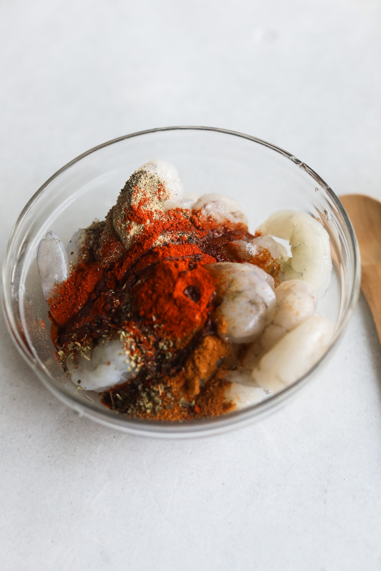 Raw shrimp in a small glass bowl with blackening seasoning. Small wooden spoon on the side of the bowl.