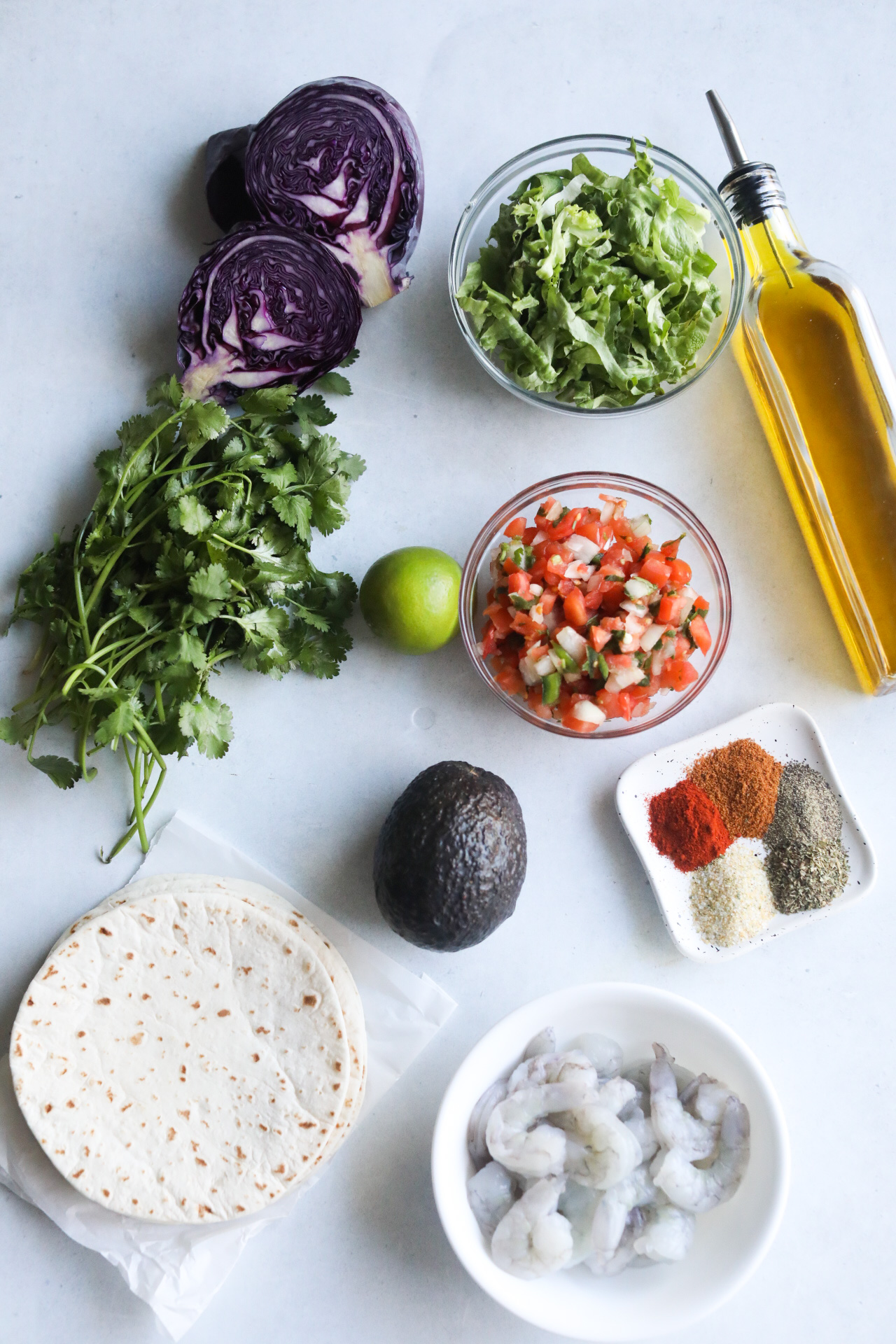 Blackened shrimp tacos ingredients on a flat lay. Red cabbage, clear bowl with shredded lettuce, cilantro, small bowl pico de Gallo, lime, avocado, blackened seasoning, tortillas, olive oil bottle and bowl of raw shrimp.