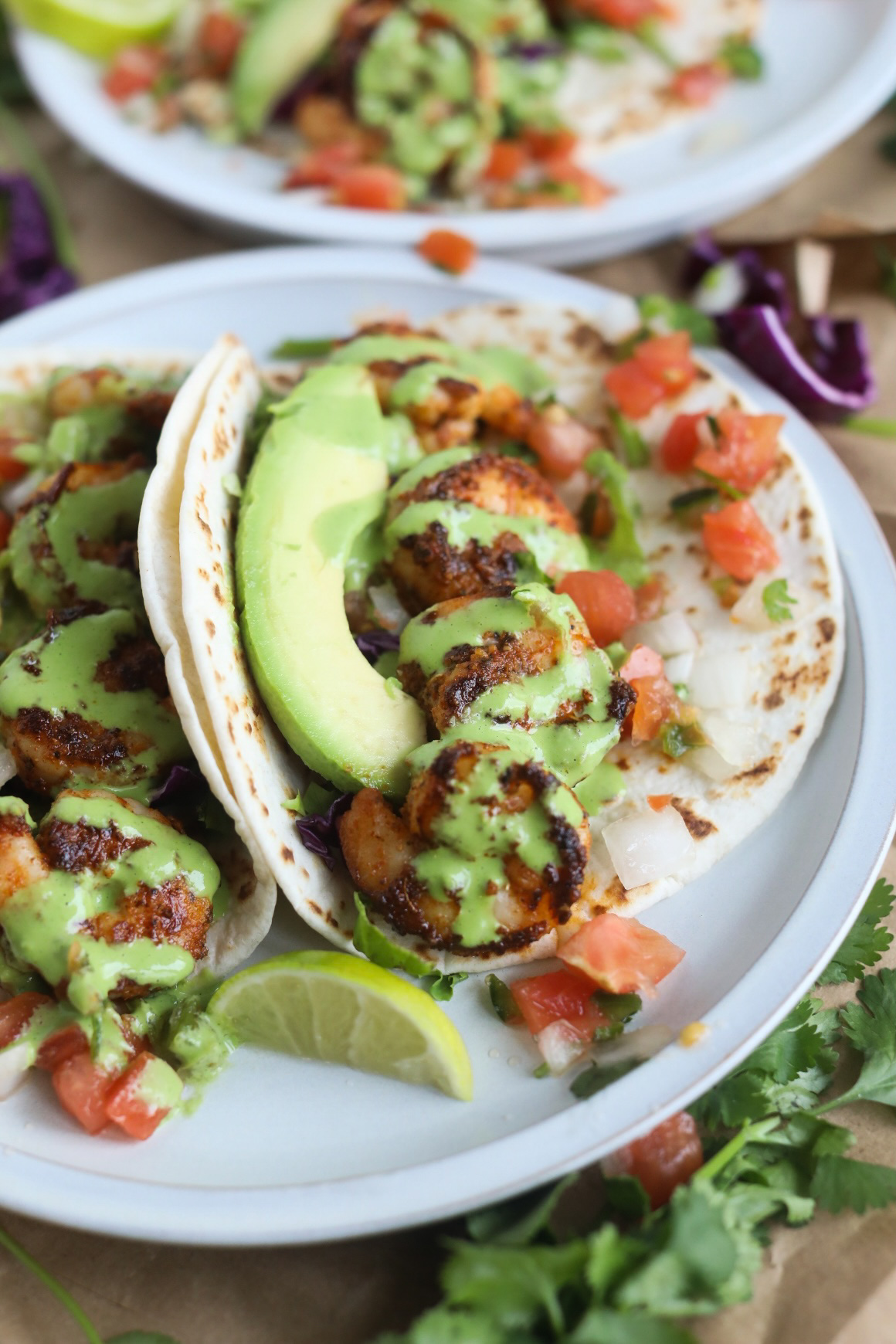 Shrimp tacos with blackened shrimp with cilantro lime sauce drizzle plated on a small grey matte plate with pico de gallo and sliced limes.