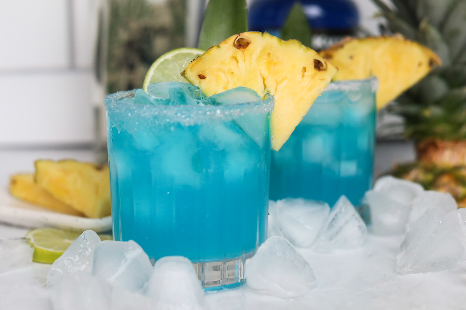 Blue margarita recipe finished. Ice cubes added for styling purposes. Margarita is bright blue and dressed with sliced pineapple, lime and pineapple stem. 