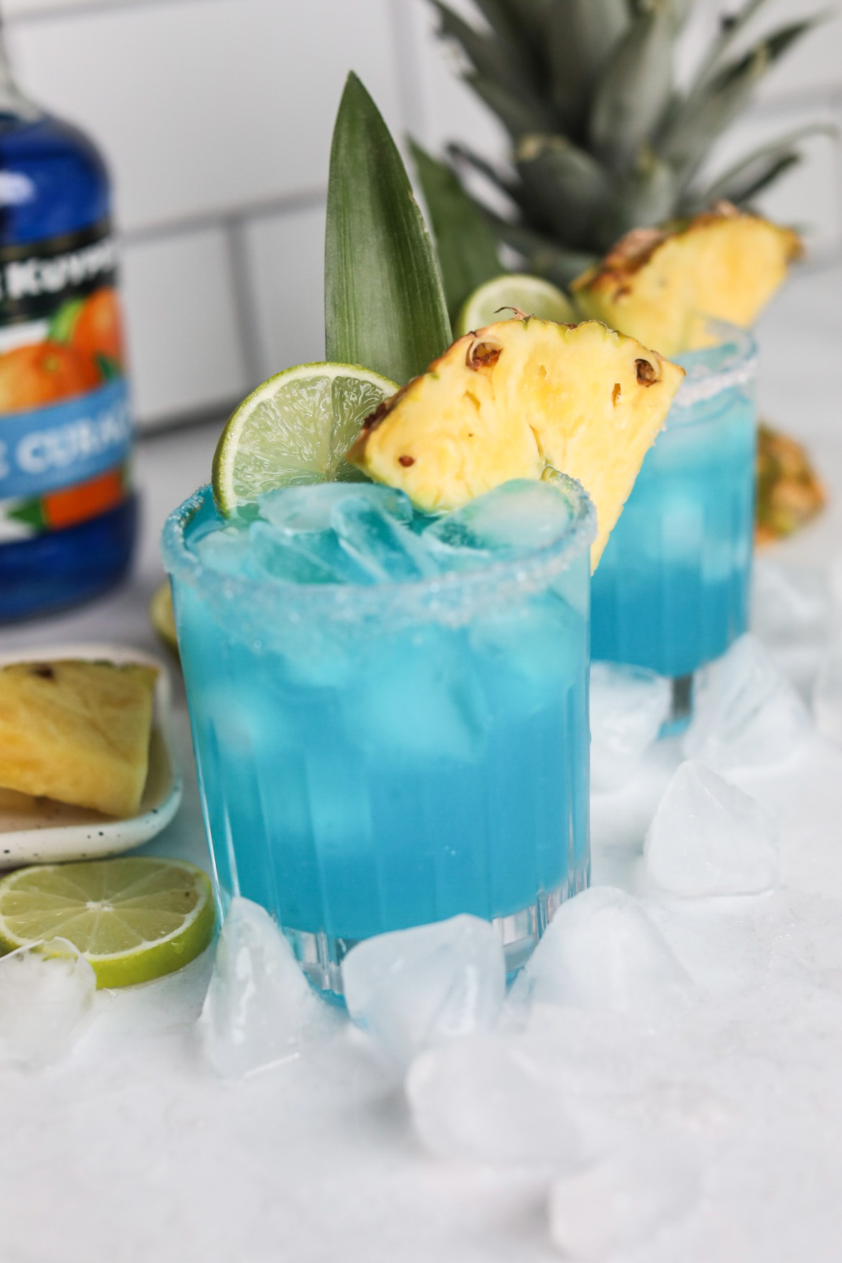 Two glasses with blue margaritas in a short glass. Glasses are dressed with pineapple slices, lime slice and pineapple stem. For styling purposes, ice cubes, sliced limes and pineapple top is added. 