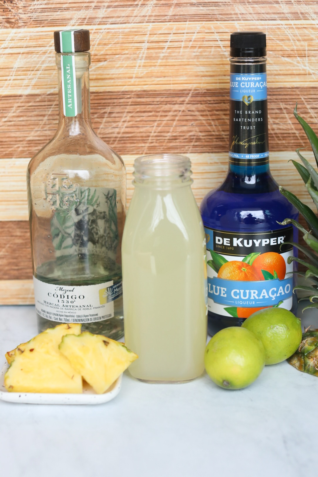 Blue Margarita Ingredients. Bottle of tequila, homemade sour mix in a glass jar and blue curaçao. Garnishes are plated on a small white plate consisting of pineapples, two whole limes and a pineapple top. 