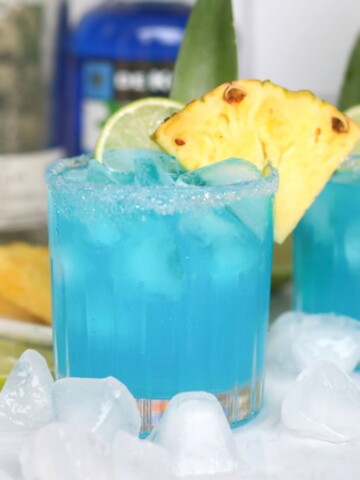Blue margarita recipe featured image with one cocktail as the focal point. Margarita is dressed with a salted rim, sliced pineapple, lime and pineapple stem.