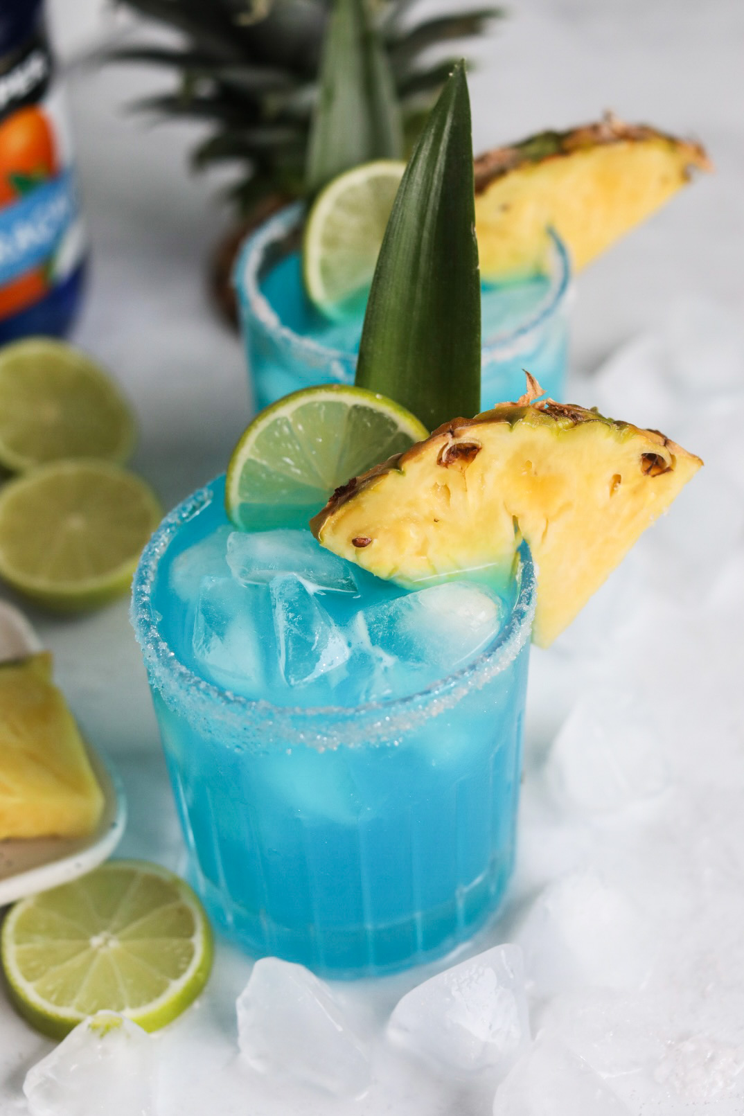 Blue margarita recipe dressed with salt, sliced pineapple, limes and pineapple stem. Ice cubes are added for styling purposes. 